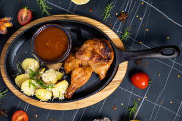 Half of a roasted chicken, on a black plate with herbs and tomatoes, on a dark concrete