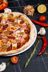 Aromatic pizza with hunting sausages, onions and tomatoes. As package design element. Pizza on a black wooden background among spices