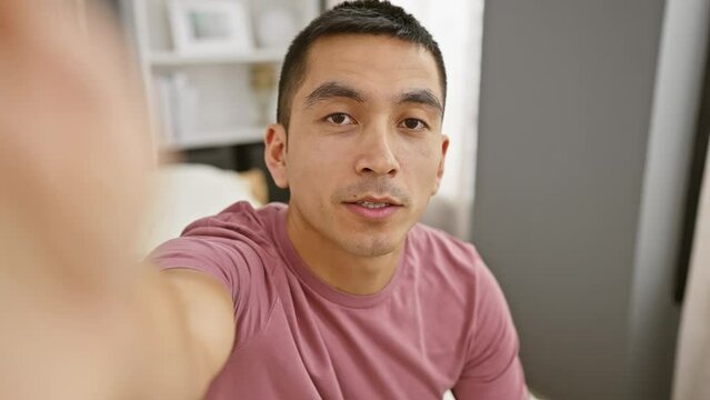 A smiling young hispanic man taking a selfie in a cozy bedroom setting, depicting a relaxed, casual atmosphere.