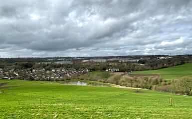 Obraz premium Landscape view from, Cliff Hollins Lane, with a large field, a lake, and distant houses, on a cloudy day near, Dewsbury, UK