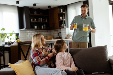 Smiling father brings a tray of orange juice to his happy family seated in a living room - 763446891