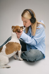 Kissing small dog Jack Russell terrier. Dog owner love and care. Blonde woman kissing nose of her pet sitting on the floor listening to music. Studio shot with gray background