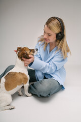 Blond woman hugging small dog Jack Russell terrier. Sitting on the floor listening to music. Blue jeans and shirt and gray background studio shot
