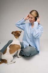 Pretty girl and pet music lover. Blonde woman sitting on the floor with black headphones listening to song cute white dog looking at her with love. Studio shot. Gray and blue colors