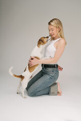 Dog kissing blonde woman pet owner. Happy laughing girl hugging small dog. Love emotions. Studio shot. Grey background