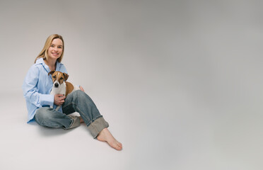 Smiling blonde woman in a blue shirt sits on the floor, the dog sitting on her laps. Funny cute friends studio portrait. Grey background. Large horizontal size banner copy empty space