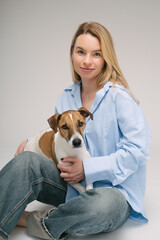 Smiling blonde woman in a blue shirt sits on the floor, the dog sitting on her laps. Funny cute friends studio portrait. Grey background. Vertical composition