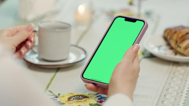Close-up Of Mobile Phone With Green Mock-up Screen In Female Hands. Woman Uses Smartphone While Resting In Cafe, Drinks Tea and Eats Croissant