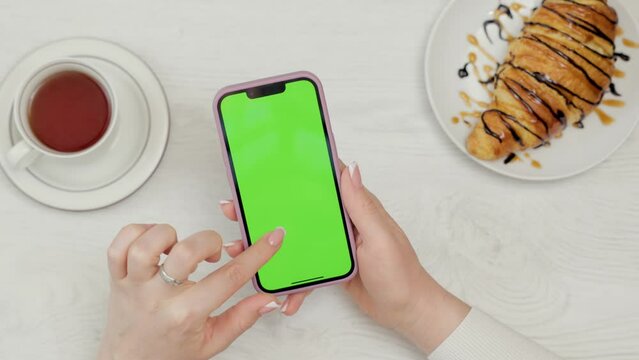 Close-up Of Mobile Phone With Green Mock-up Screen In Female Hands. Woman Uses Smartphone While Resting In Cafe, Drinks Tea and Eats Croissant