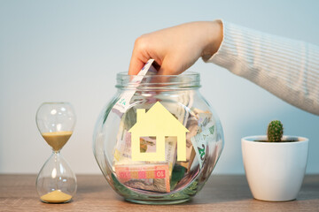 Taking saved money for a house or flat in Ukraine. Female hand taking out hryvnia banknotes out of jar with house symbol, buying a new house