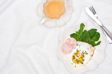 Healthy breakfast with fried egg, chicken sausage, spinach leaves, pumpkin seeds, green tea on white linen background