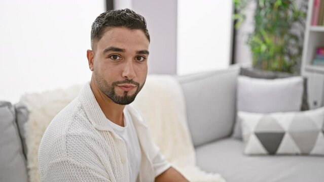 Confident young arab man in his natural pose sitting at home on the sofa, a handsome chap with a cheeky smile, looking off side with a relaxed vibe.