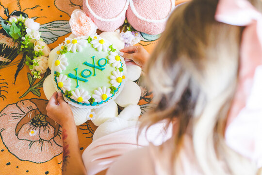Cute XOXO Flower Cake with Daisy Flowers on Gothic Floral Blanket Setup, Cake Held by Blonde Model Wearing Bow, Creative Photo of Anniversary Celebration, Dating, or Valentine's Day