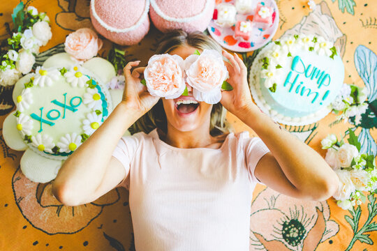 Cute and Cozy Photograph of Model Lying with Fake Peony Flowers Over Her Eyes Surrounded by Pillows, Flowers, and Cakes. Funny Floral Cakes Say: XOXO and Dump Him