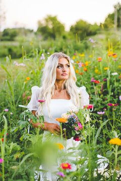 Beautiful Blonde Model Sitting in Zinnia Wildflower Garden, Colorful Golden Hour Sunset Bridal Portrait of Bridal Model with Loose Hair Wearing Modern Wedding Dress with Puffy Sleeves