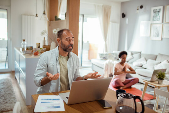 Man working on laptop with woman in background doing yoga at home