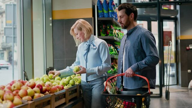 Charming female selecting apples from wooden crates in the produce section and putting them in shopping basket holding by bearded husband. Romantic atmosphere in supermarket and time together.