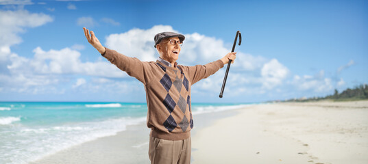 Elderly man with a cane spreading arms on a sandy beach - obrazy, fototapety, plakaty
