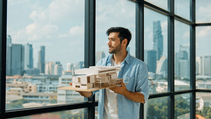 Skilled smiling caucasian engineer turns house model left and right to check mistake point. Professional manager inspect at architectural model while standing near window. Design concept. Tracery - obrazy, fototapety, plakaty