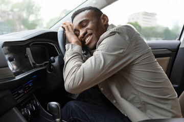 Delighted African American man hugging the steering wheel of his new car, closing his eyes and...