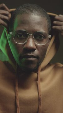 Vertical chest up portrait of young serious Black male programmer pulling up hood and looking at camera while green programming code appearing on black background