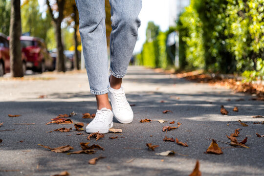 Cropped photo of woman legs in white sneakers going on sidewalk with dry leaves fall season outside