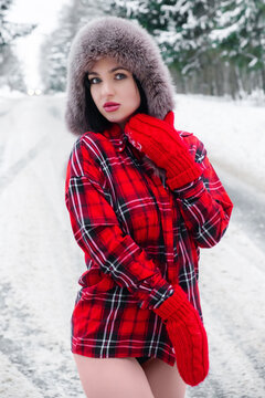 pretty girl in a red checkered shirt, earflap hat, knitted red mittens with bare legs stands looking at the camera against the backdrop of a snowy forest road