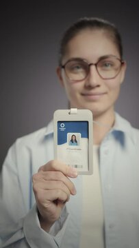 Vertical medium slowmo portrait of young Caucasian female IT coordinator showing her ID badge at camera standing on grey studio background