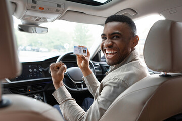  Happy young african man showing his driver's license while driving a car - obrazy, fototapety, plakaty