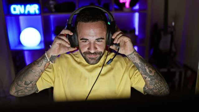Smiling bearded man with tattoos wearing headphones in a colorful illuminated gaming room at night.