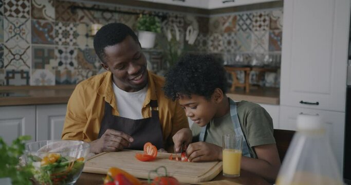 Slow motion of little boy making salad enjoying cookery sitting at table with loving father in kitchen. Happy childhood and nutrition concept.