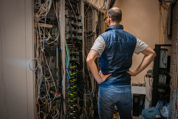 A technician looks at a rack of server equipment, viewed from the back. A man with his hands on his belt is thinking about how to solve a problem in a data center.