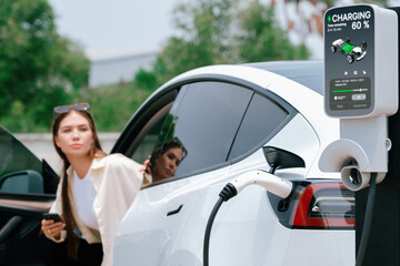 Young woman use smartphone to pay for electricity at public EV car charging station green city...