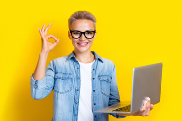 Photo portrait of pretty young girl specs hold netbook show okey symbol wear trendy jeans outfit isolated on yellow color background - obrazy, fototapety, plakaty