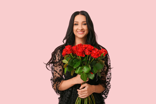 Beautiful young woman with dark wavy hair in black dress holding roses on pink background