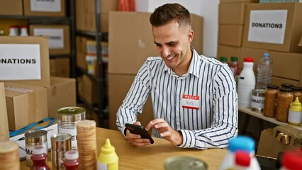A young man named alex is smiling while using a smartphone in a donation center with various food items and boxes in the background.
