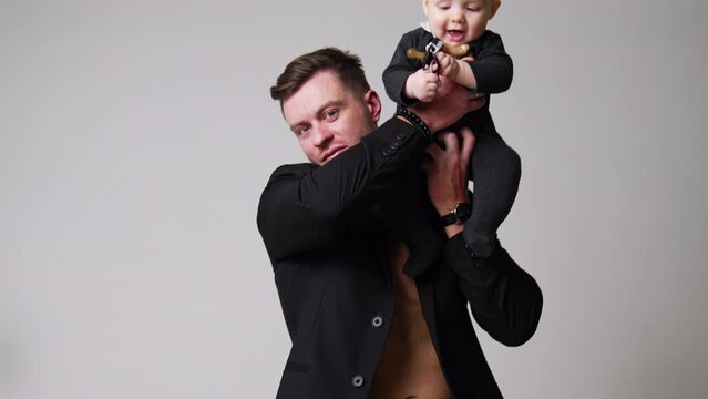 Handsome man in black jacket sits holding a baby. Father puts his lovely little child on the shoulder. White backdrop.