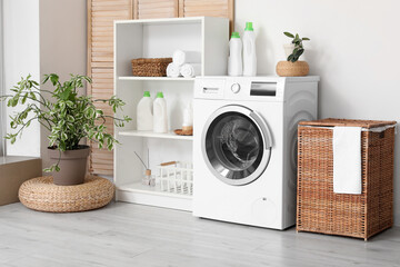 Modern washing machine with basket, shelving unit and dressing screen near white wall. Interior of home laundry room - obrazy, fototapety, plakaty