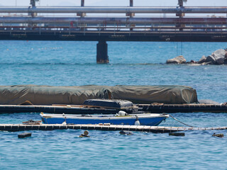 A boat with blue and white colors is anchored near a massive inflated barrier in calm sea. In the...