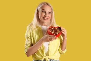 Mature woman with box of heart-shaped chocolate candies on yellow background