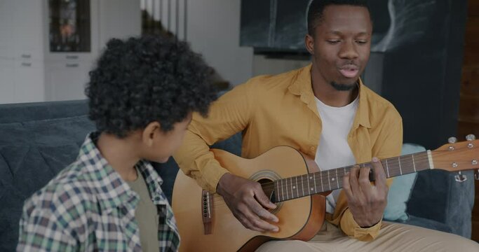 African American man father playing the guitar and singing with child son enjoying leisure time at home. Creative youth and family relationship concept.