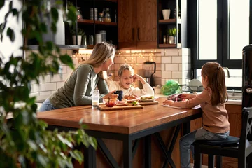 Fotobehang Mother and two daughters share the kitchen table, with mom and the elder sister working on homework together while the younger one joyfully doodles. A delightful morning of togetherness. © DusanJelicic