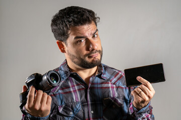 Hombre sosteniendo un celular y una cámara digital en expresión de duda y comparación