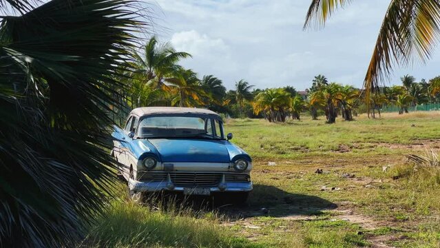 VARADERO, CUBA - DECEMBER 2, 2024: An old American car standing under a palm tree, illuminated by the sun. A retro car. Blue classic American car on the beach. 4К