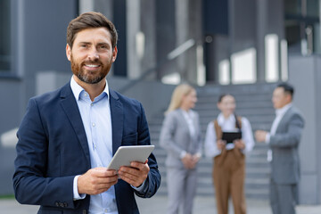 Smiling business professional using a tablet with blurred colleagues in the background, depicting leadership and technology integration in a modern corporate environment.