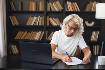 Middle aged senior woman using laptop computer writing notes at workplace. Focused mature old businesswoman work at office making written records doing online work. Mature boss professional worker - obrazy, fototapety, plakaty