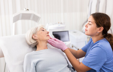 Glad old woman having consultation about face aesthetic with a doctor lying on clinical chair in the cabinet