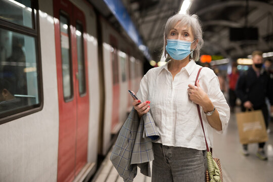 Senior European woman in face mask standing beside arrived train in subway station.