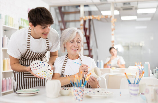 Positive young guy assisting enthusiastic senior woman painting ceramic mug in pottery class, showing plate with paint samples. Intergenerational communication and creative hobby concept