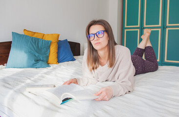 Young woman in eyeglasses lying down on the bed and studying with reading books at home
